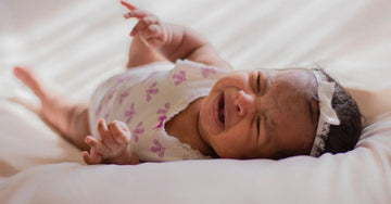 A crying newborn baby lying on a soft bed, expressing the need for comfort and physical contact. The image illustrates the importance of promptly responding to a baby’s cries to support their emotional security and healthy development.
