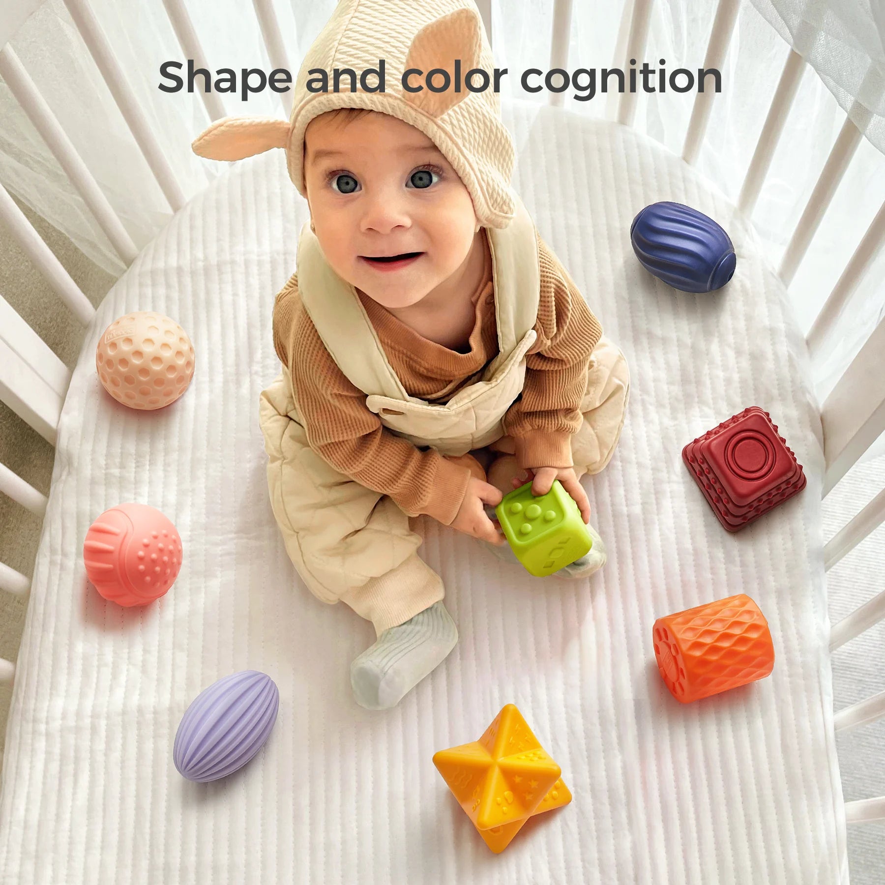 A baby dressed in a beige romper and animal-ear hat sits in a white crib, surrounded by colorful textured sensory blocks in various shapes including spheres, cubes, and stars. The baby holds a green cube and looks up with a smile. The text "Shape and color cognition" appears above, emphasizing the toy's educational value for early learning.