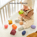 A baby dressed in a beige quilted romper and ear-flap hat sits in a crib, intently examining a green sensory cube. Surrounding the baby are various colorful sensory balls and blocks with different textures and shapes, supporting tactile exploration and early learning. The text 