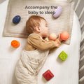 A baby sleeps peacefully in a crib while holding two textured sensory balls—one peach-colored and one orange. Several other colorful sensory blocks and balls are scattered around on the mattress and pillow. The baby is dressed in a beige quilted romper, and the text “Accompany the baby to sleep” appears at the top, emphasizing comfort and play.