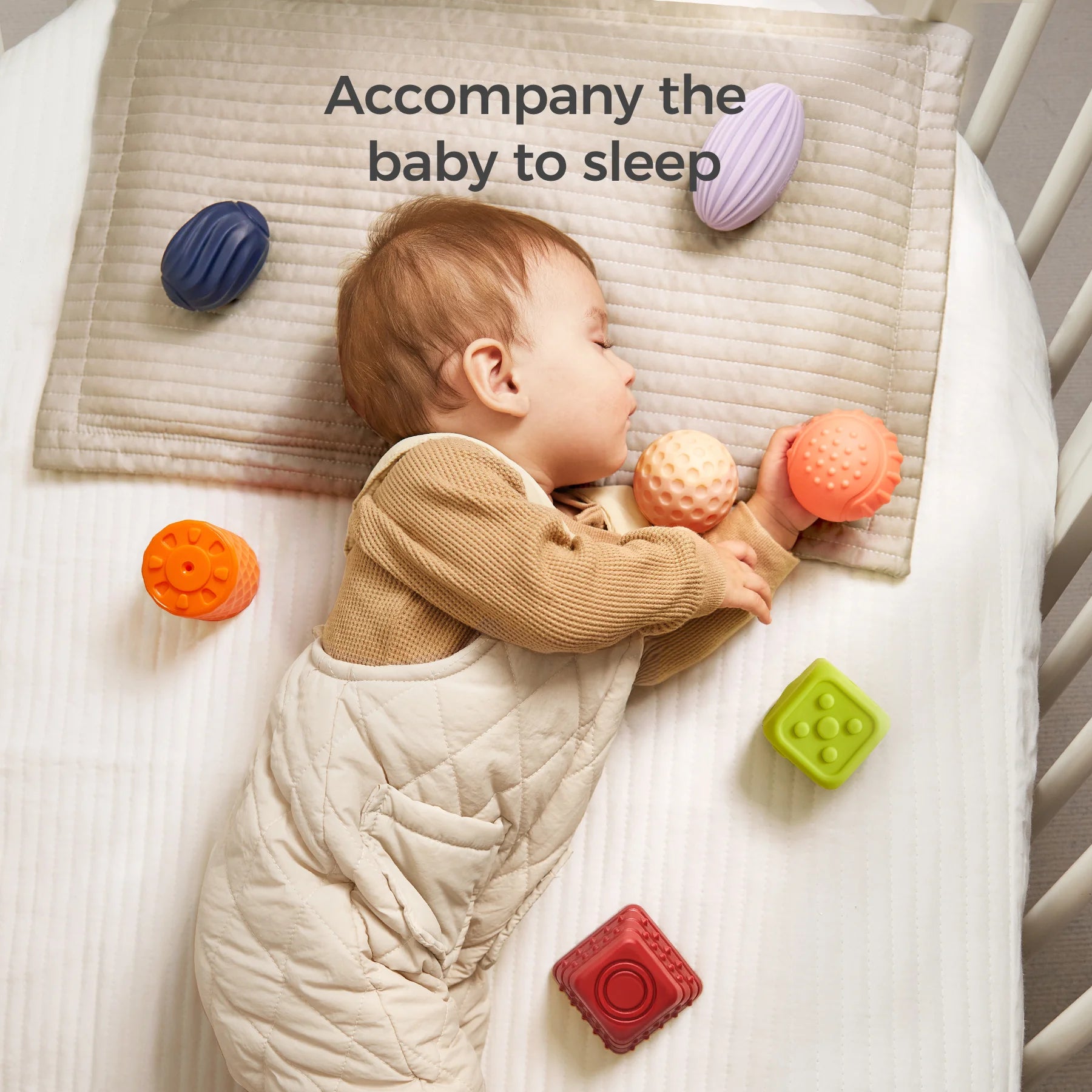 A baby sleeps peacefully in a crib while holding two textured sensory balls—one peach-colored and one orange. Several other colorful sensory blocks and balls are scattered around on the mattress and pillow. The baby is dressed in a beige quilted romper, and the text “Accompany the baby to sleep” appears at the top, emphasizing comfort and play.