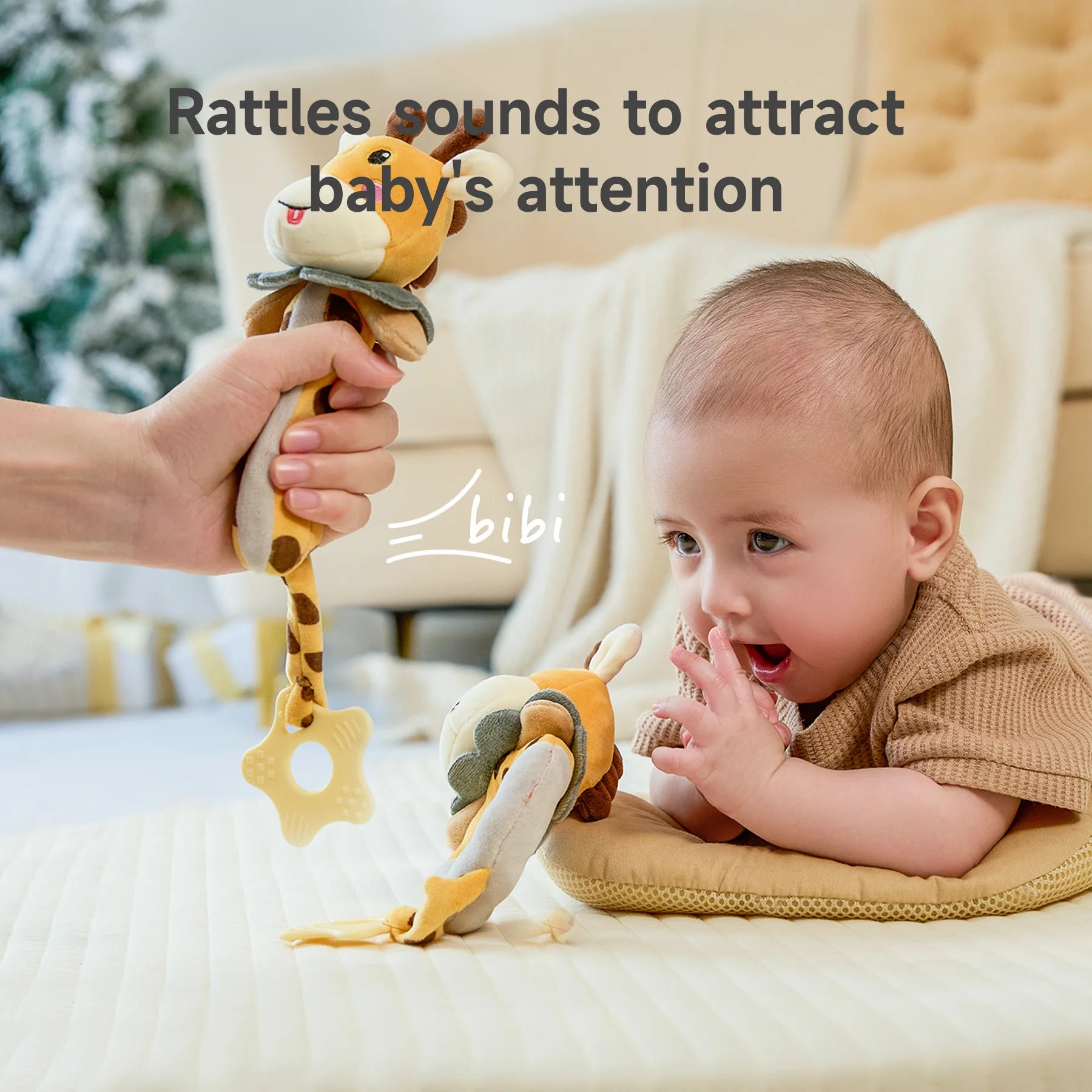 A baby lies on their tummy, excitedly looking at a plush giraffe rattle being held by an adult. The giraffe toy has a teething ring at the bottom, and another giraffe rattle is placed near the baby. The text “Rattles sounds to attract baby’s attention” is displayed at the top.