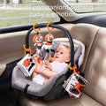 Baby sitting in a gray car seat, reaching toward black, white, and orange hanging soft toys and cloth books attached to the handle with plastic clips. The plush toys include animal characters and textured teething accessories. Text overlay says: “Travel companion – Soothing baby’s emotion.” The background shows a blurred view outside the car window.