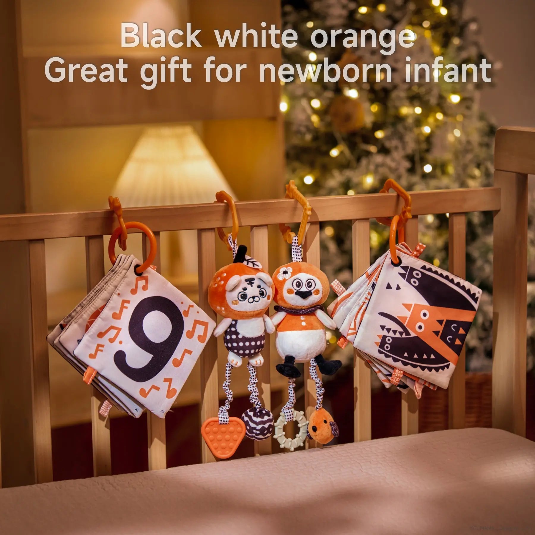 High-contrast black, white, and orange baby toy set displayed on a crib rail, featuring two plush animal toys with hanging textured teethers and two soft fabric books with numbers and bold patterns. Text overlay reads “Black white orange – Great gift for newborn infant,” with a cozy, warmly lit holiday background.