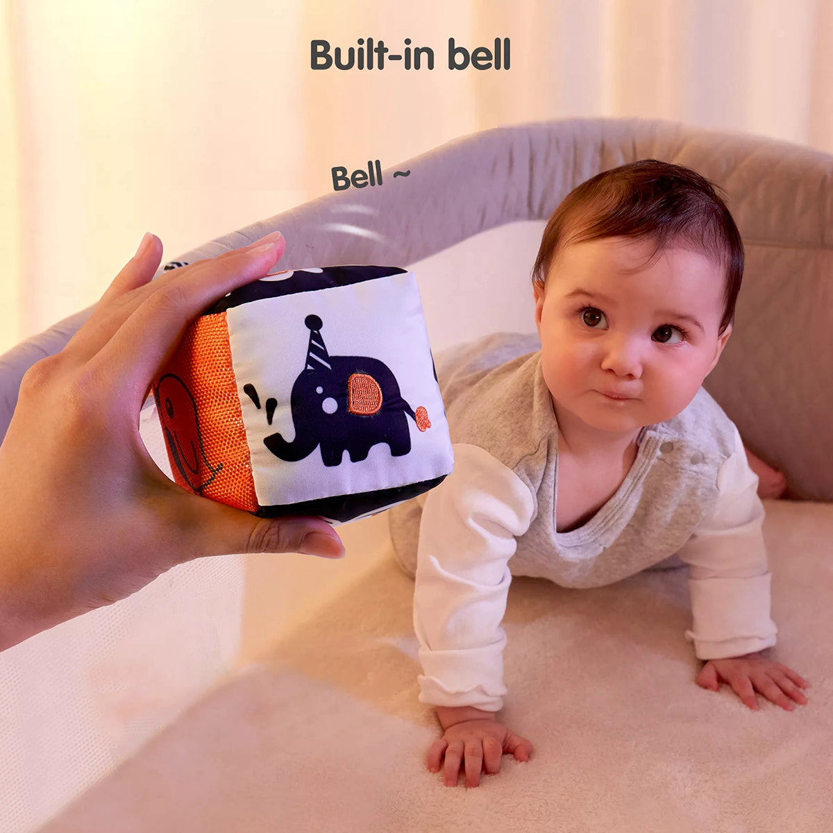 A baby on all fours looking at a soft cube toy with a built-in bell, held by an adult. The cube features high-contrast animal designs, including an elephant, and produces a gentle ringing sound to capture the baby's attention and stimulate auditory development.