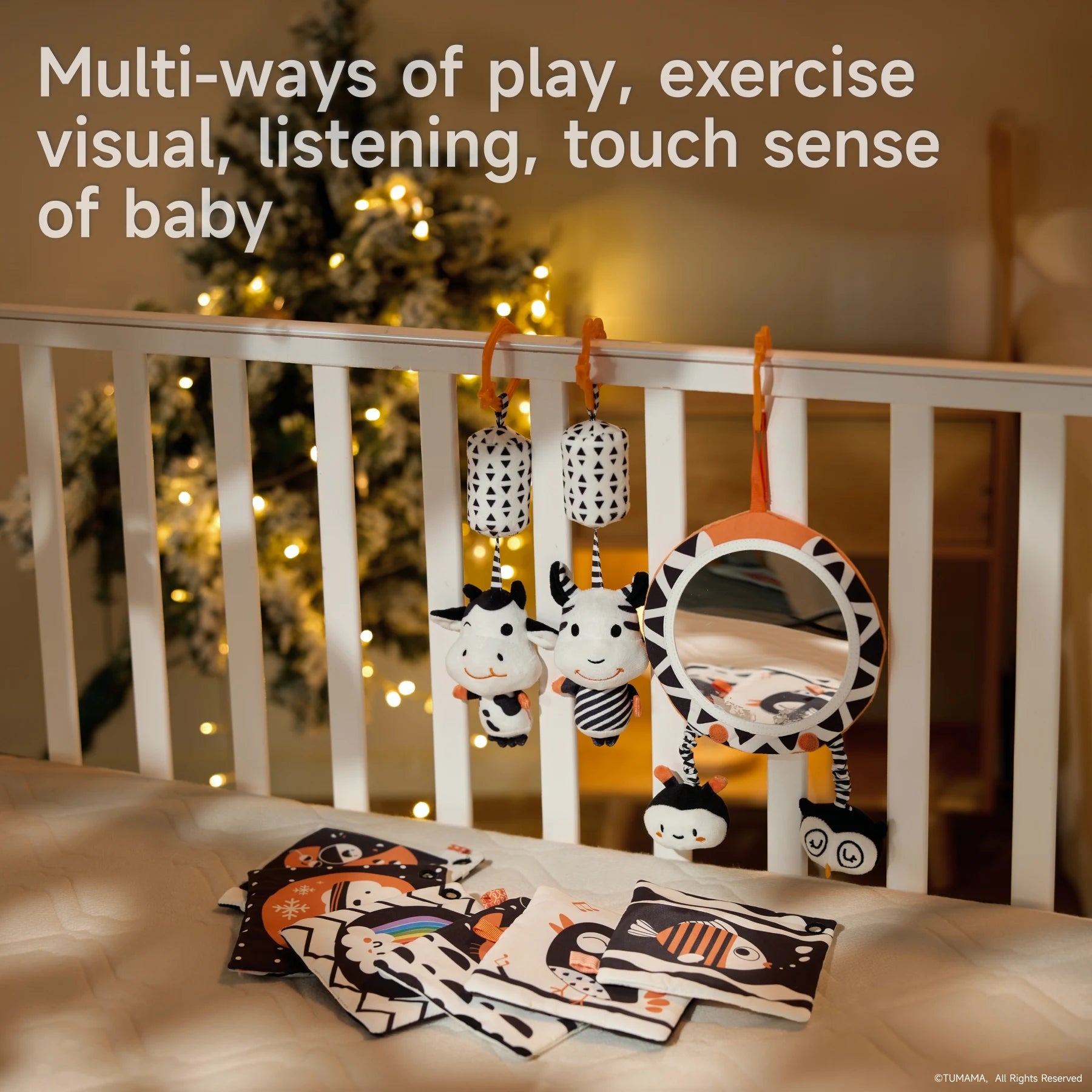Baby crib decorated with high-contrast black and white toys, including two plush cows with rattles, a circular mirror with owl and bee plushies, and several soft cloth books spread on the mattress. A Christmas tree with warm lights is in the background. Caption reads “Multi-ways of play, exercise visual, listening, touch sense of baby.”