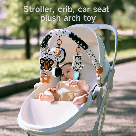 Baby lying in a white stroller outdoors with a black and white plush arch toy attached overhead. The toy features colorful hanging plush characters including a dog, owl, and butterfly, designed for use with strollers, cribs, and car seats.