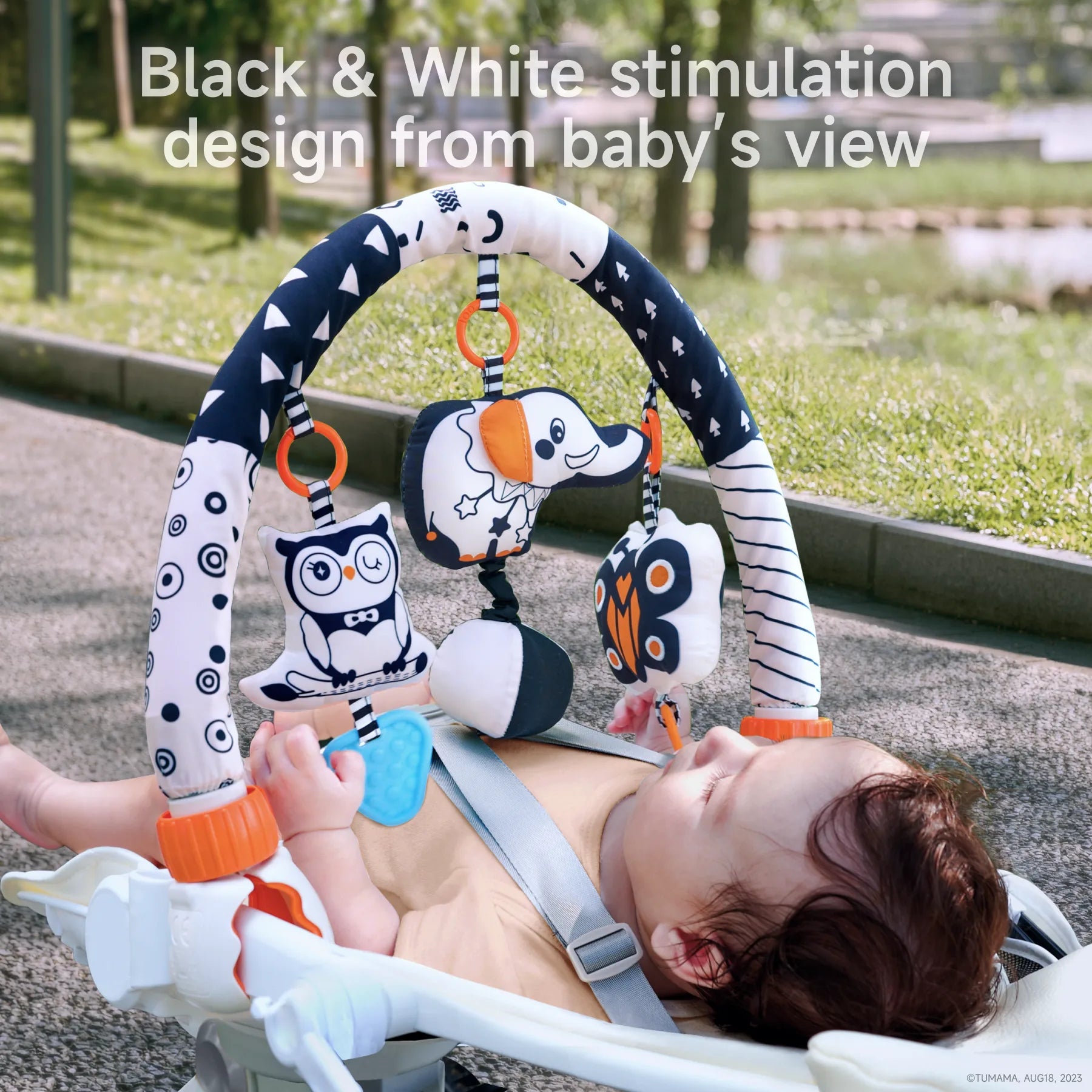 Baby lying in a stroller and gazing up at a plush arch toy featuring black-and-white patterns with orange accents, designed for visual stimulation. Hanging toys include an owl, dog, butterfly, and textured teether. The arch is positioned for optimal viewing and sensory engagement from the baby’s perspective.
