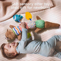A joyful baby in light blue ribbed clothing lies on a cream-colored quilted surface, happily playing with a soft sensory otter toy. The plush toy features a caterpillar-like design with a brown otter head with cute facial features at one end and a tail at the other. Between them are five colorful textured segments in different patterns: striped, pink with circles, multicolored squares, and green with dots. Attached to the toy are a blue whale teether and a yellow leaf-shaped teether. Text overlay at the top