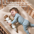 A smiling baby in light blue ribbed pajamas lies on a cream-colored crib mattress, hugging the soft otter sensory toy that doubles as a measuring tool. The toy is stretched out alongside the baby, showing its full length with measurement indicators clearly visible on the underside. The plush toy features a brown otter head at one end and colorful segments between, with measurement markings ranging up to 40cm as indicated by text at the bottom of the image. The blue whale teether is visible with a zigzag lin