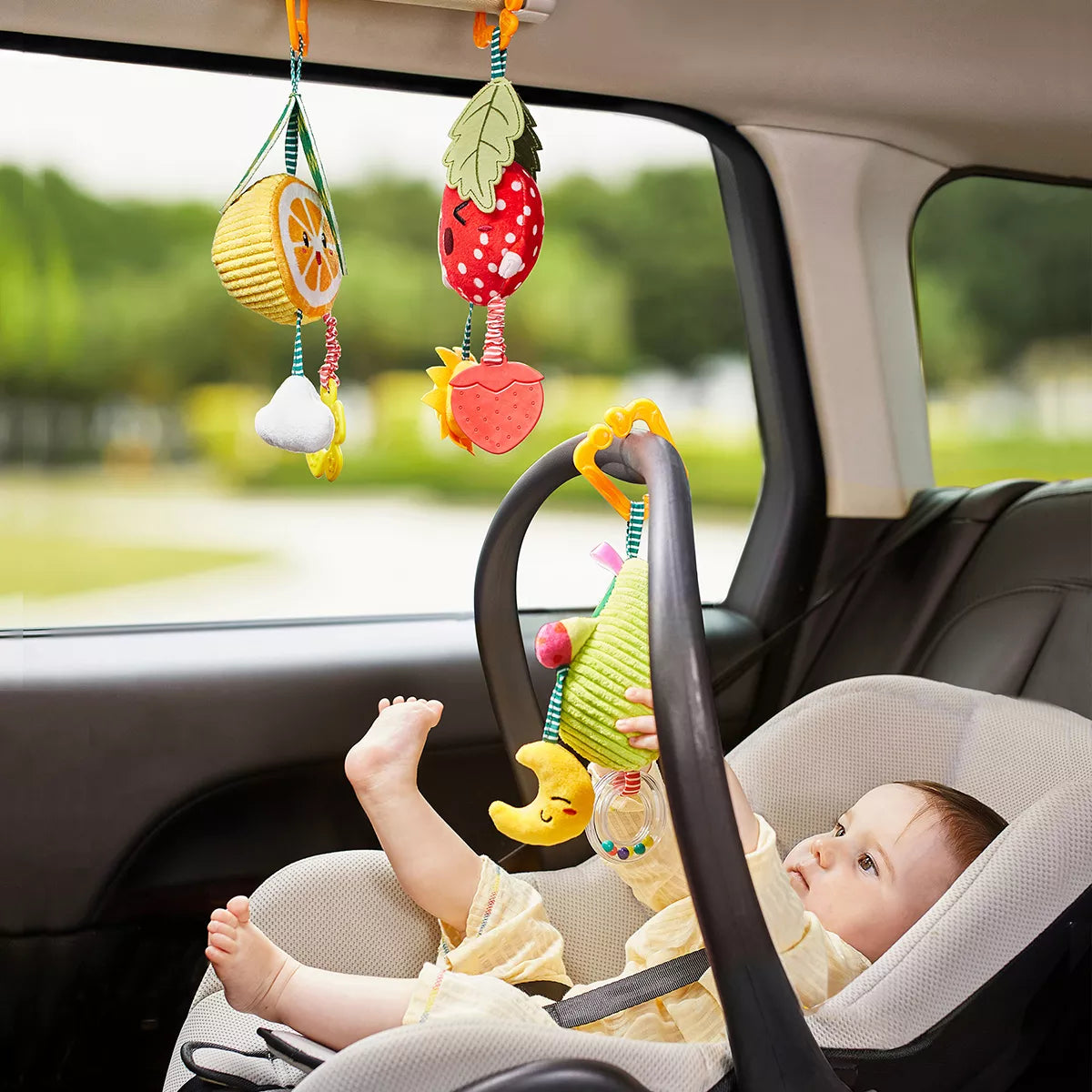 A baby seated in a car seat playing with colorful plush fruit-shaped hanging rattle toys attached to the car interior. The toys, including avocado, orange, and strawberry designs, feature soft textures and dangling accessories, keeping the baby entertained and stimulated during travel.