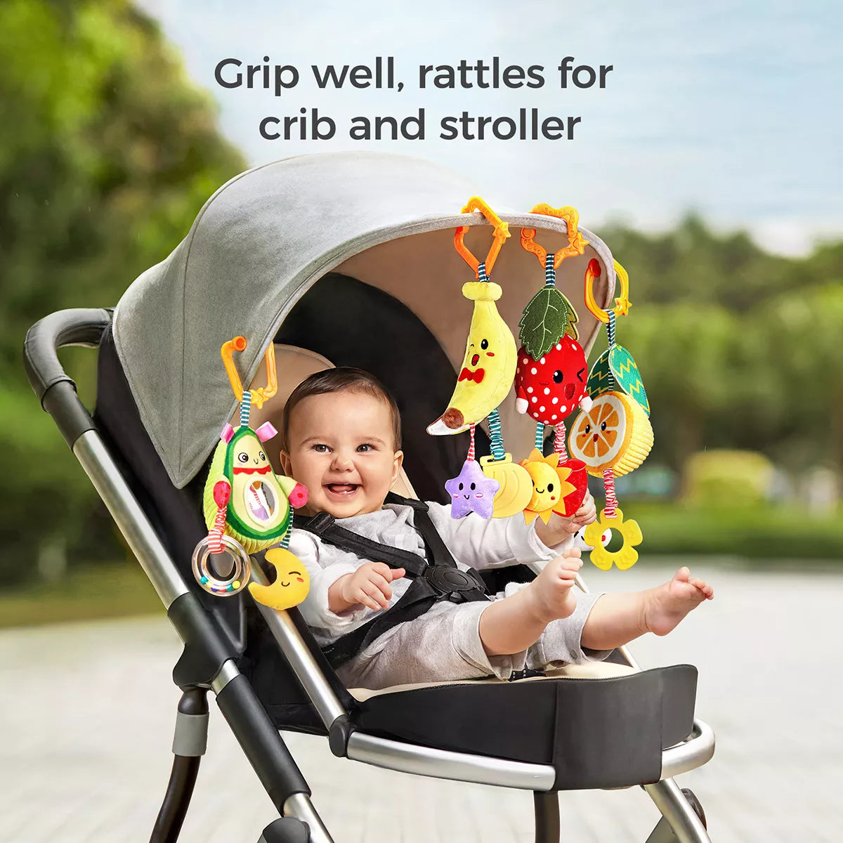 A happy baby sitting in a stroller, playing with colorful plush fruit-shaped hanging rattle toys including avocado, banana, strawberry, and orange. The toys are attached to the stroller canopy with orange clips, making them ideal for both crib and stroller play to entertain and stimulate babies.
