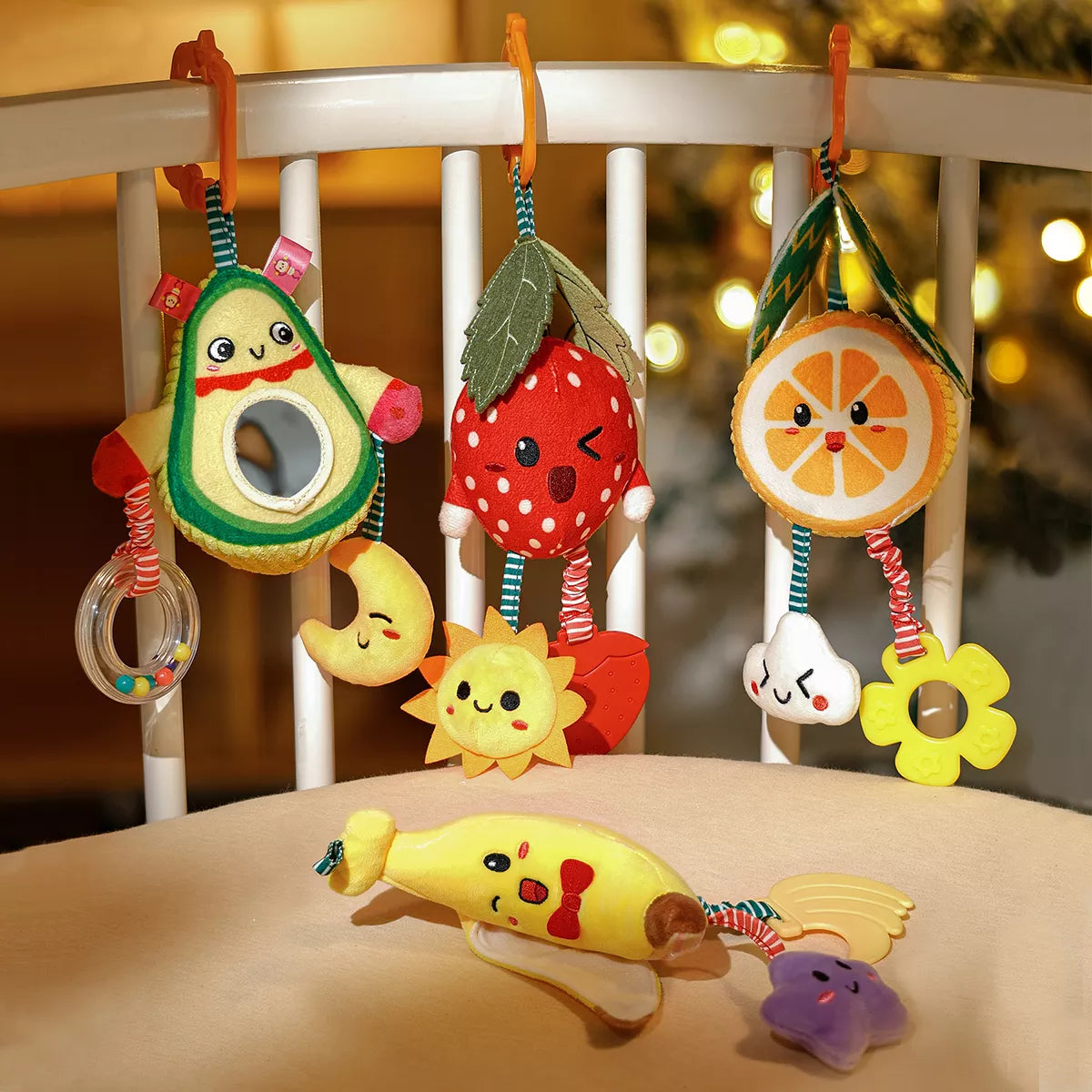 Set of plush fruit-shaped baby rattle toys including avocado, strawberry, orange, and banana, hanging on a crib rail with orange clips. Each toy features cute embroidered faces, mirrors, or teething accessories like stars, moons, and flowers. Designed to entertain and soothe babies while encouraging sensory development.