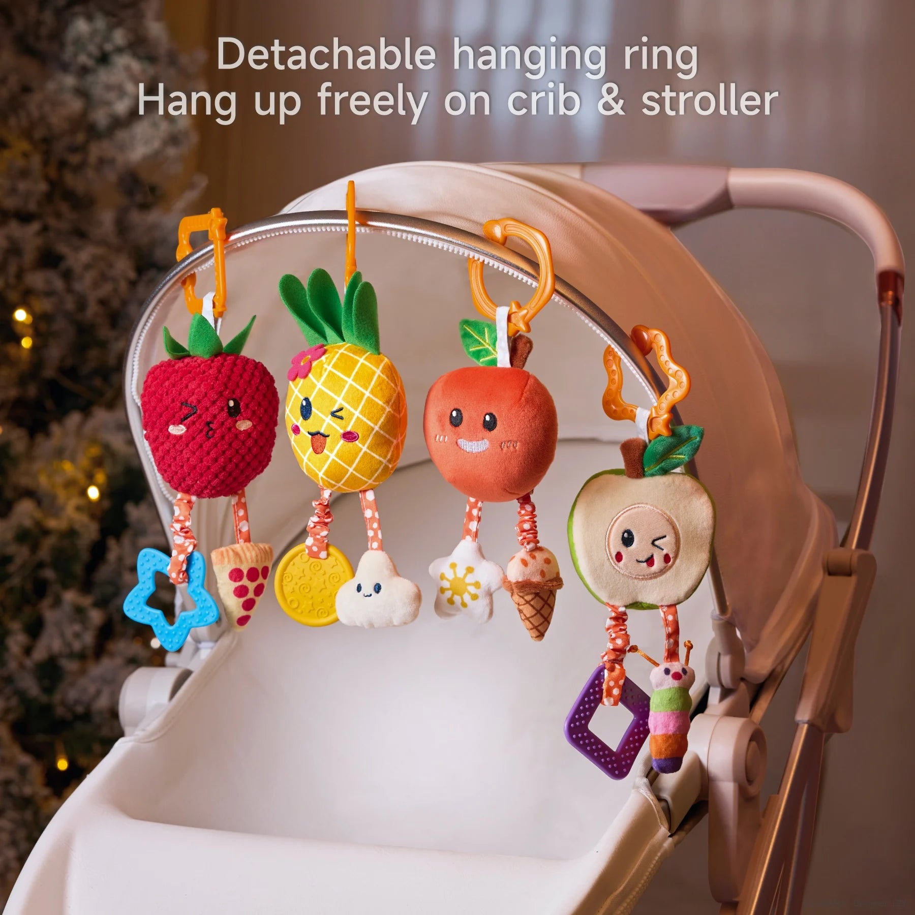 Four colorful plush fruit rattle toys — strawberry, pineapple, orange, and apple — are hanging from a stroller canopy using detachable orange rings. Each toy features soft fabric textures, cheerful embroidered faces, and dangling accessories like teethers and rattles. The background includes warm indoor lighting and a softly lit Christmas tree. Overlay text reads: “Detachable hanging ring. Hang up freely on crib & stroller.”