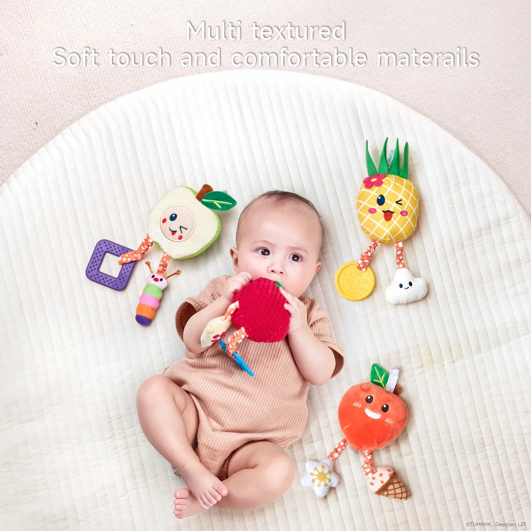A baby lies on a soft, cream-colored playmat, surrounded by four colorful plush fruit toys — apple, strawberry, pineapple, and orange — each with dangling textured accessories. The baby is holding and mouthing the red strawberry toy. The image highlights the toys' multi-textured, soft-touch materials. Text on the image reads: “Multi textured. Soft touch and comfortable materials.”