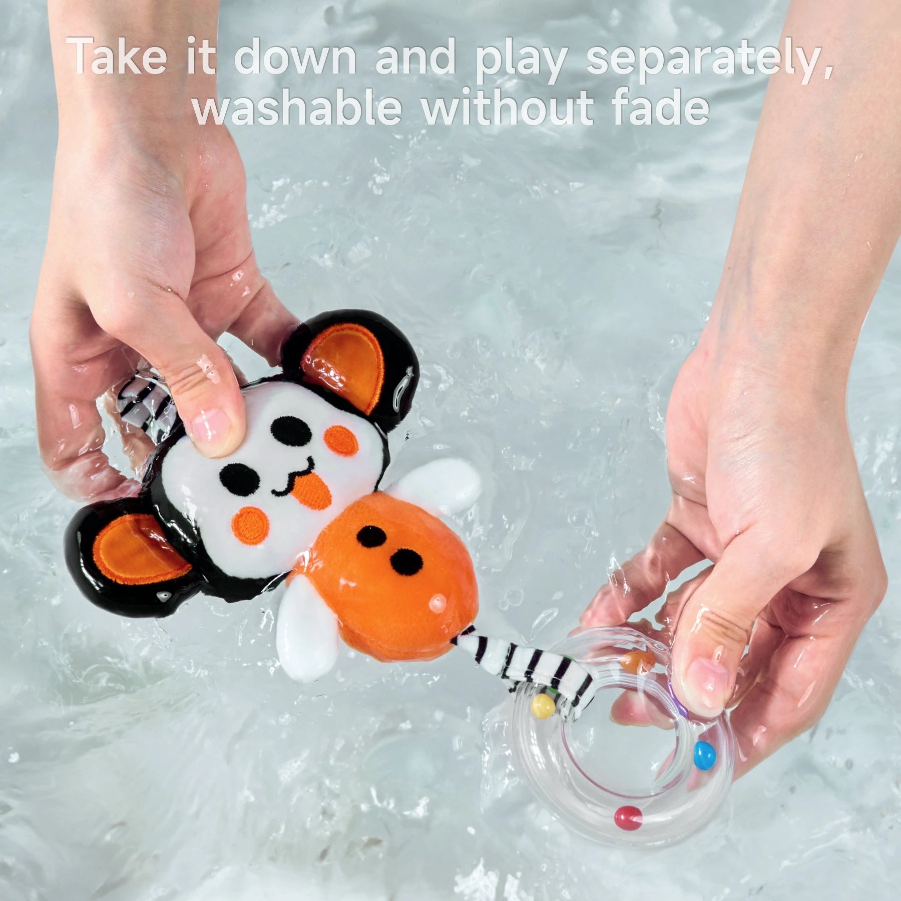 Hands washing a plush monkey toy with a clear rattle ring in a basin of water, demonstrating that the toy is detachable, playable separately, and washable without fading.