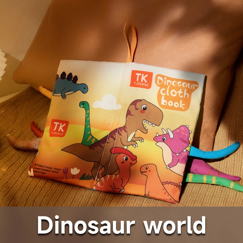 Colorful Dinosaur Cloth Baby Book from TK Tumama displayed on a brown cushion. The cover features playful cartoon dinosaurs including a T-Rex, Triceratops, and Brachiosaurus, with various soft, textured tails extending from the sides. 
