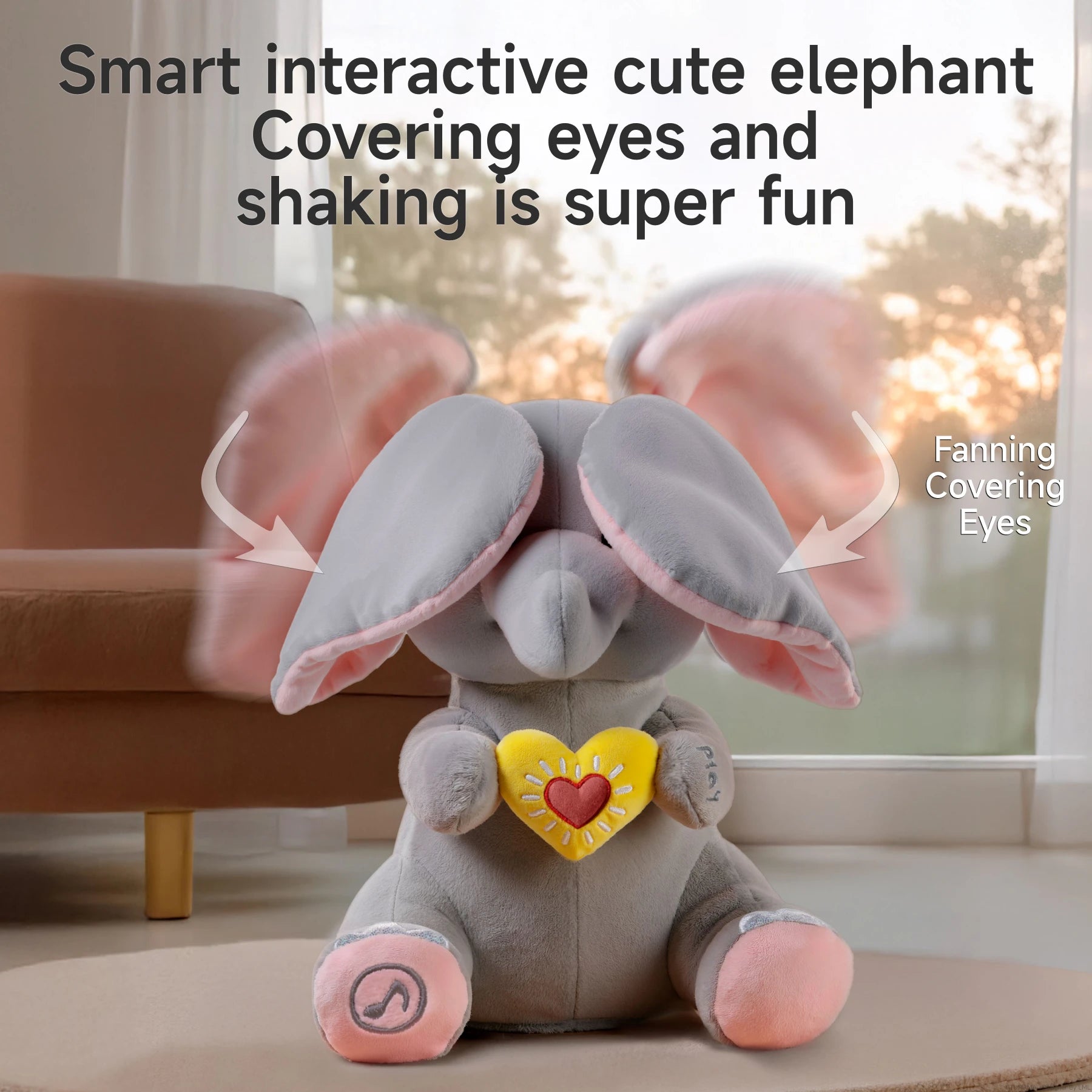 Plush elephant toy with flapping pink ears covering its eyes in a peek-a-boo motion. Text reads: “Smart interactive cute elephant — Covering eyes and shaking is super fun,” with arrows labeling the ear movements as “Fanning” and “Covering Eyes.” Sunlight shines through the window in the background.