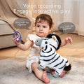 Toddler sitting on a soft carpet holding an interactive zebra plush toy with black stripes, gray hooves, and a black mane. The child is also holding a purple paw-shaped voice recording remote connected by an orange cord. Text on the image highlights 'Voice recording' and 'Engage interactive play' with speech bubbles saying 'Sing with me together'.