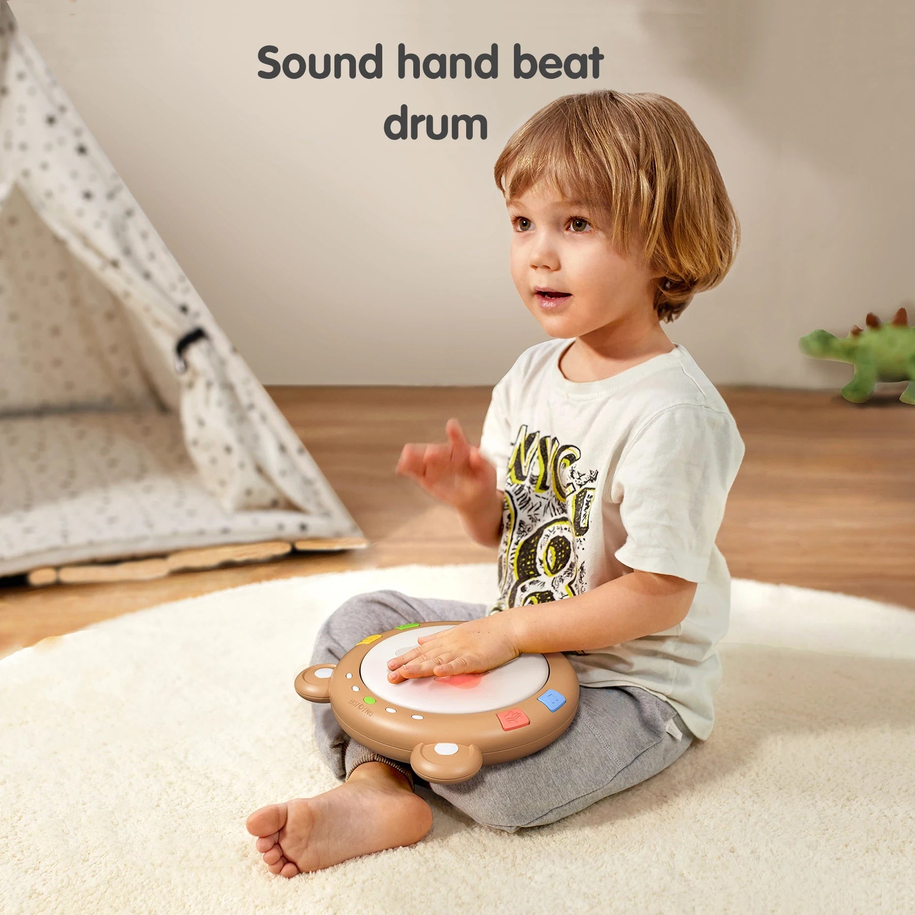A toddler sits cross-legged on a round rug, tapping a bear-shaped light-up drum toy placed on their lap. The toy's central surface glows red when touched, and colorful buttons line the edges. A soft indoor play area with a tent and toy dinosaur is in the background. The text "Sound hand beat drum" appears above, highlighting the toy’s musical interaction.