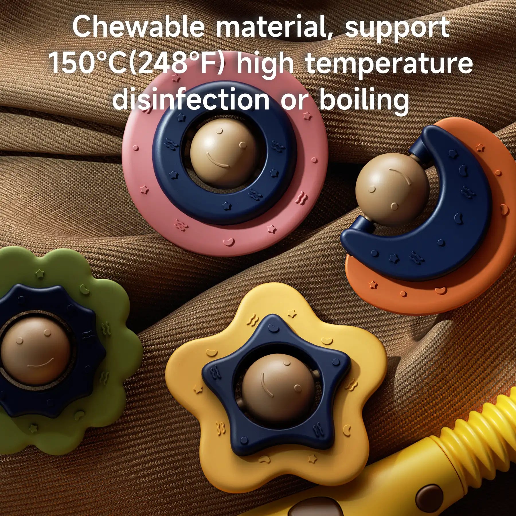 Close-up of four colorful baby crib mobile toys placed on a brown fabric background. The toys are shaped like flowers, stars, and moons, each with a smiling round face in the center. They are made of chewable material. White overlay text reads: “Chewable material, support 150°C (248°F) high temperature disinfection or boiling.”