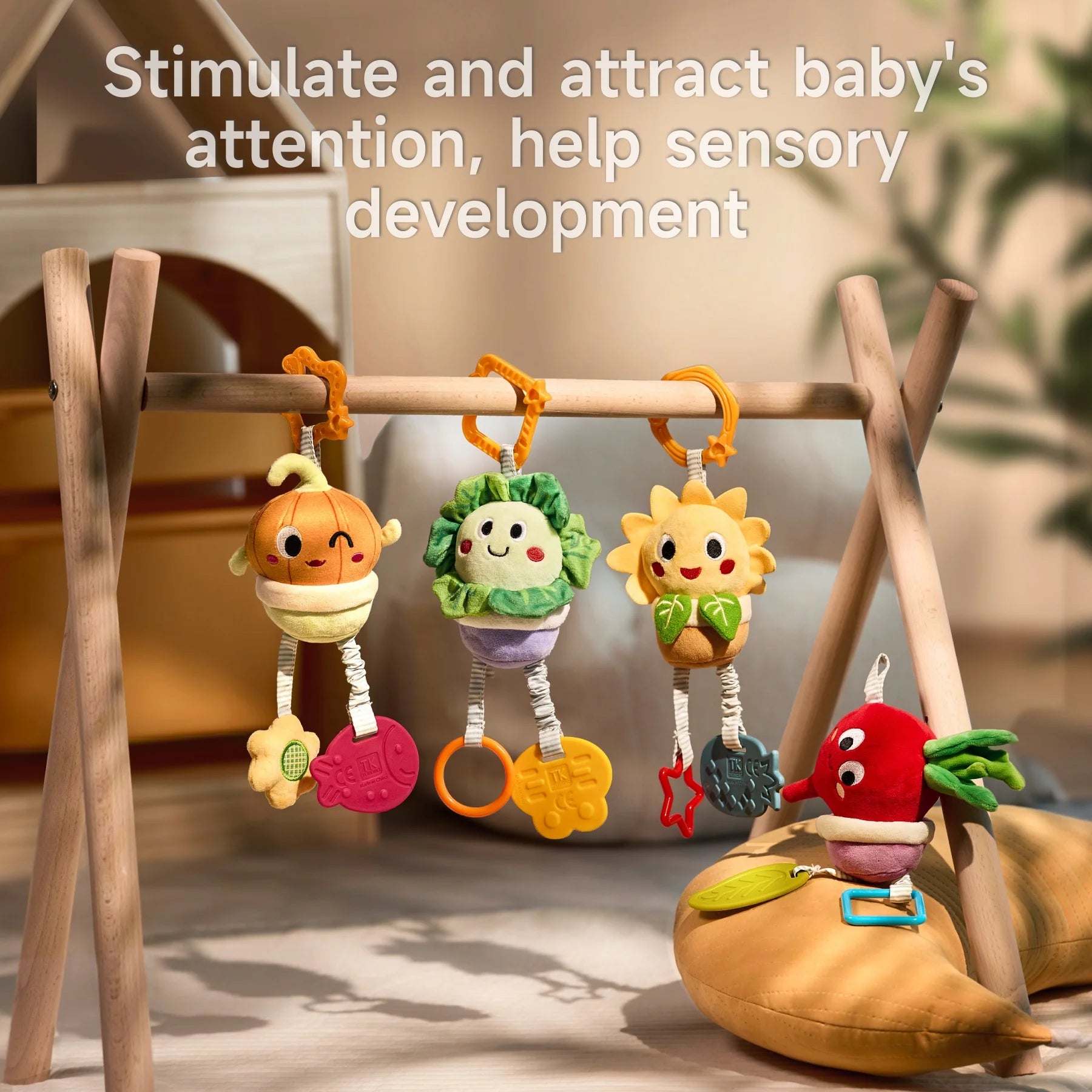 Four colorful plush stroller toys shaped like potted plants—a smiling onion, cabbage, sunflower, and beet—hang from a wooden play gym frame with dangling textured teethers; one beet toy lies on a cushioned leaf below, set in a cozy, softly lit nursery environment.