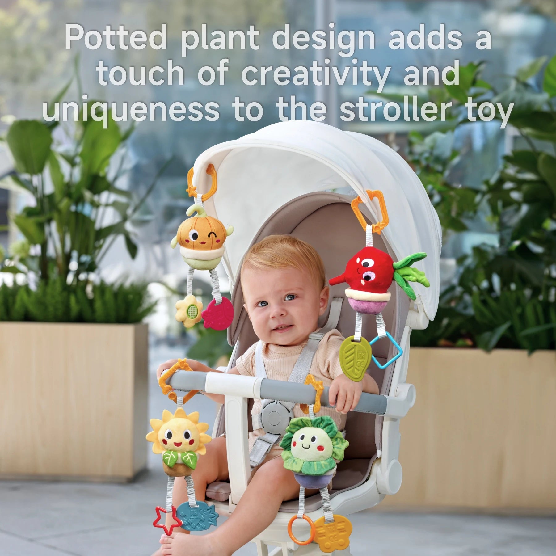 Baby seated in a stroller playing with a colorful stroller toy featuring soft plush potted plant characters like a sunflower, beetroot, onion, and cabbage, with dangling teethers and sensory elements; background includes green indoor plants and large windows.