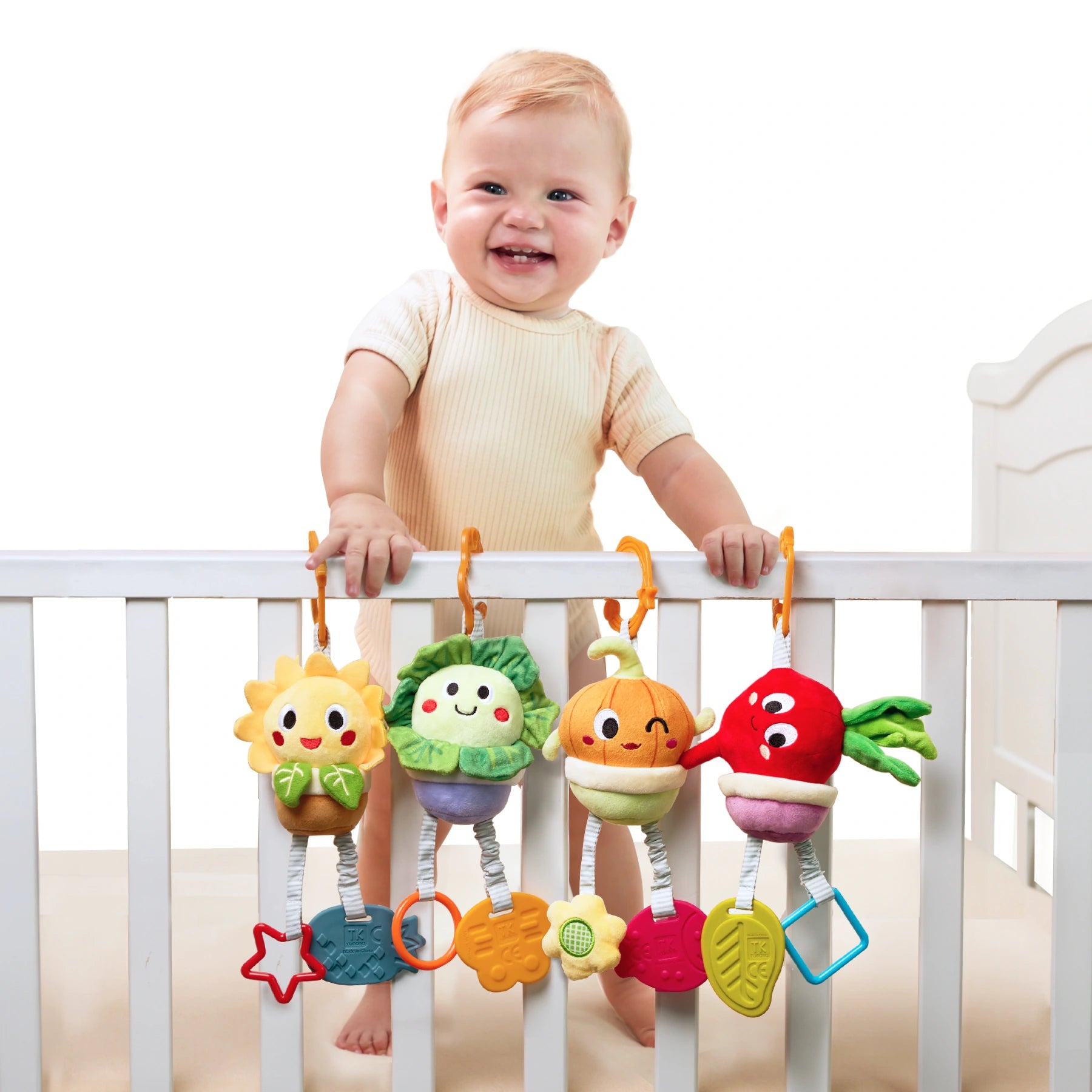 Smiling baby standing in a white crib, holding onto the railing with four colorful plush vegetable toys hanging from orange hooks. Each toy has a different design, including a sunflower, lettuce, onion, and beetroot, all with cheerful faces and textured teether shapes attached below.