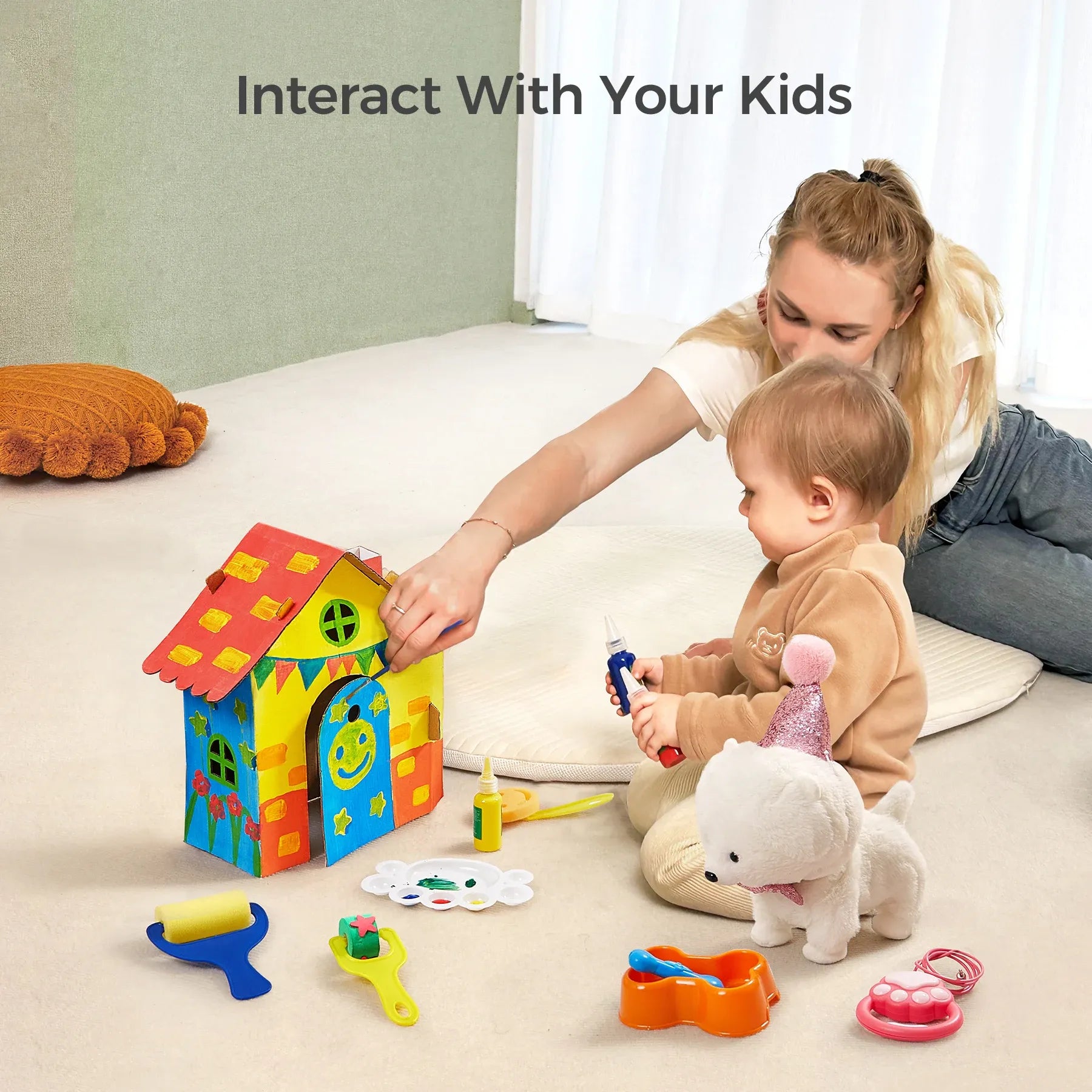 Toddler engaging in a creative activity with a white plush toy dog wearing a pink glittery party hat. The child is holding painting tools and sitting beside a brightly painted wooden doghouse, surrounded by paint bottles, a palette, and a feeding bowl with a toy bone. DIY painting set encourages artistic expression and imaginative play.