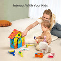 Toddler engaging in a creative activity with a white plush toy dog wearing a pink glittery party hat. The child is holding painting tools and sitting beside a brightly painted wooden doghouse, surrounded by paint bottles, a palette, and a feeding bowl with a toy bone. DIY painting set encourages artistic expression and imaginative play.