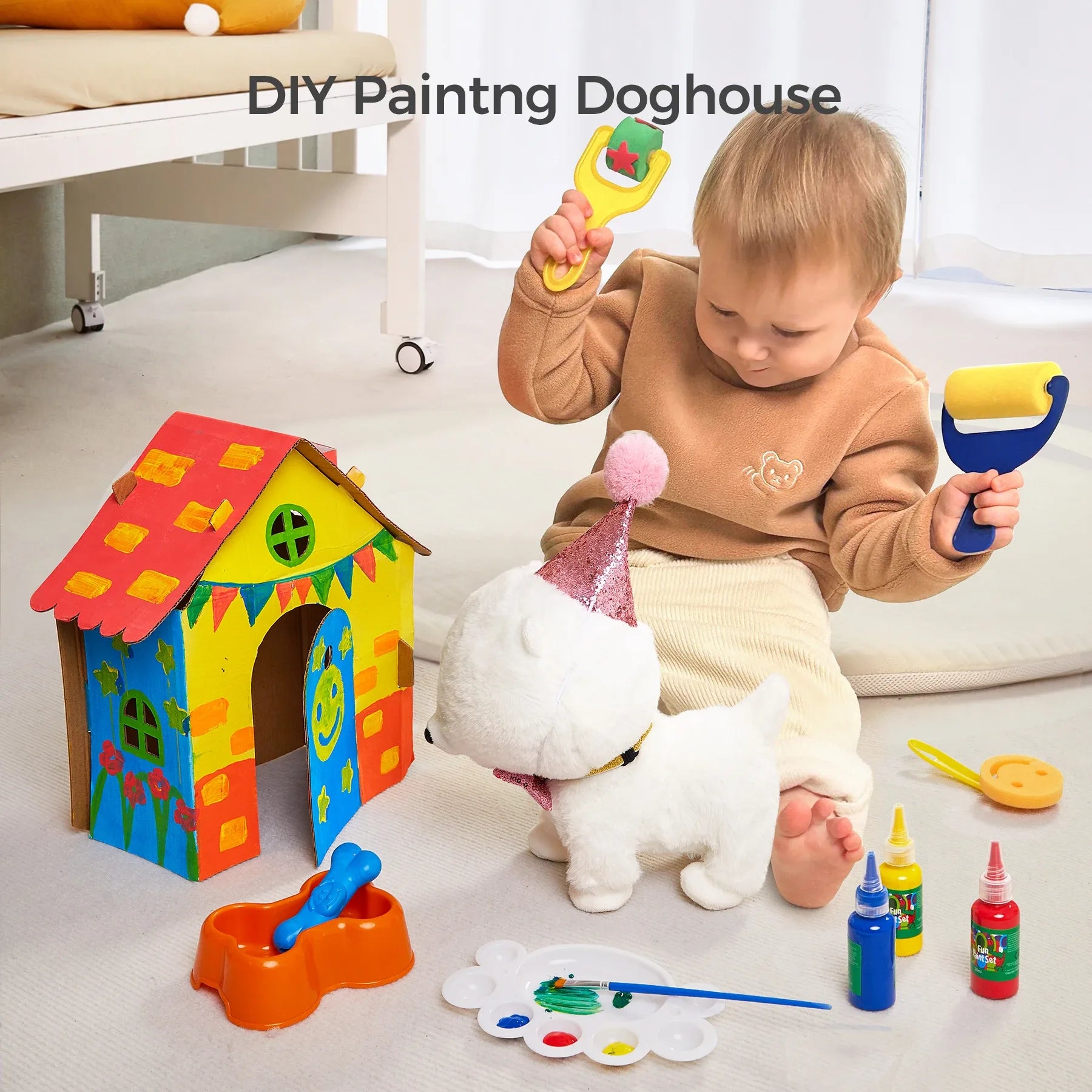 Toddler playing on the floor with a fluffy white interactive toy dog wearing a party hat, surrounded by a colorful DIY painted doghouse, paint bottles, a palette, and toy painting tools. The child is holding a paint roller and brush, engaging in creative pretend play.