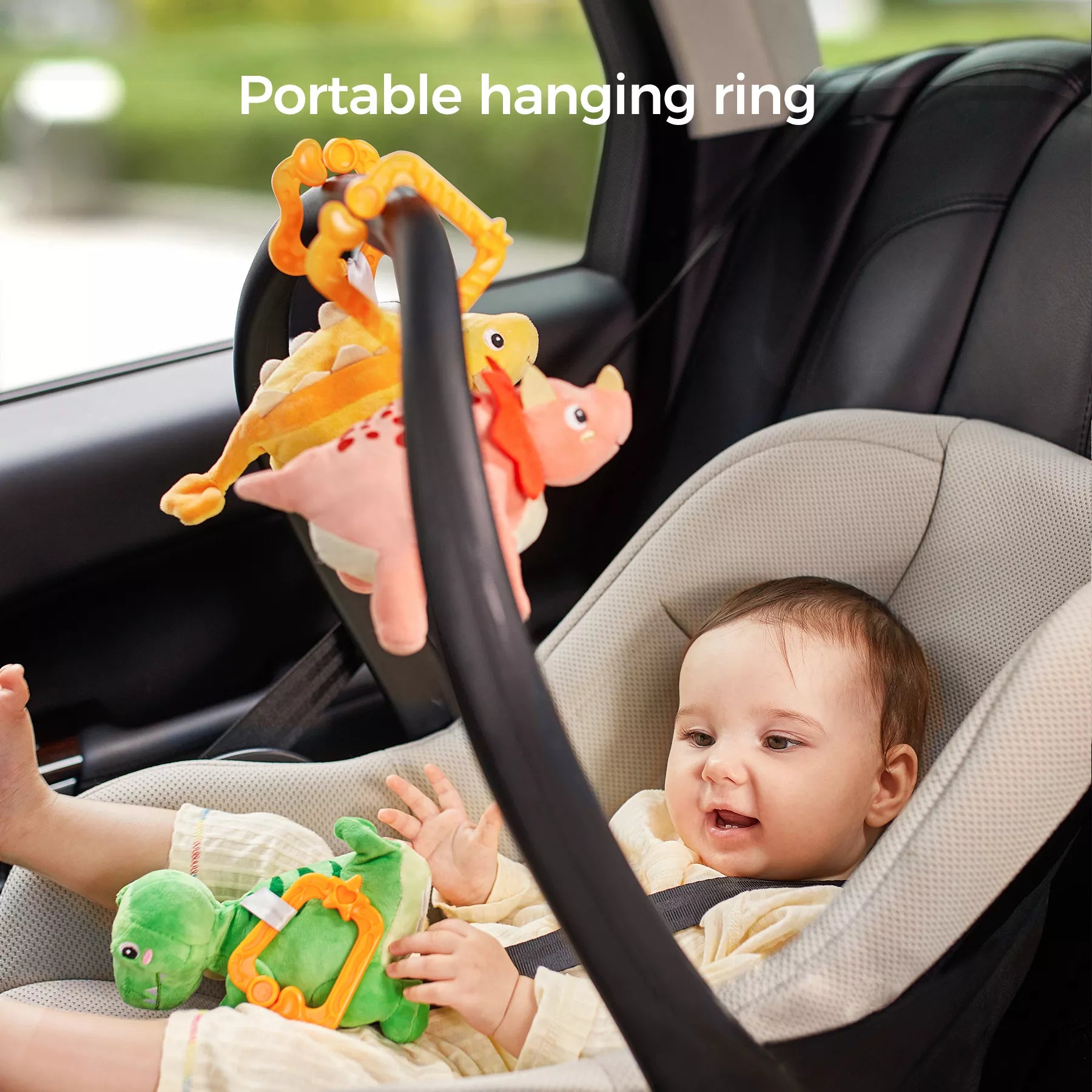Infant in a rear-facing car seat reaching for a green reversible plush dinosaur attached to an orange portable hanging ring, with yellow and pink dinosaur pals hanging above