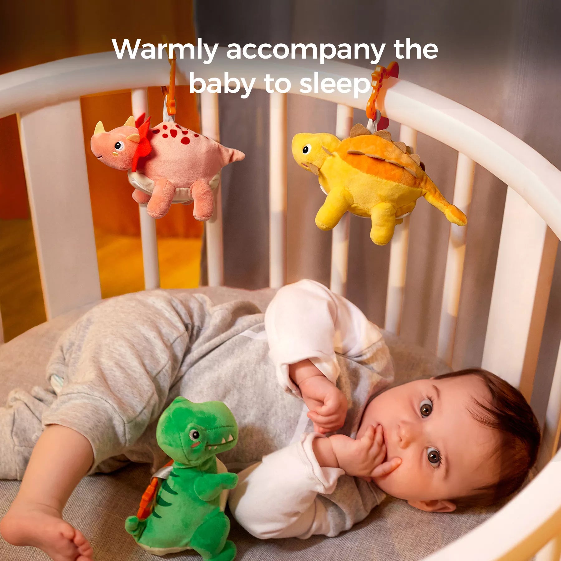 Baby lying in a white crib, reaching for a green reversible plush dinosaur toy attached to an orange hanging clip, while yellow and pink dinosaurs hang abov
