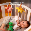 Baby lying in a white crib, reaching for a green reversible plush dinosaur toy attached to an orange hanging clip, while yellow and pink dinosaurs hang abov