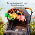 Three reversible plush dinosaur hanging toys—a green T‑rex, yellow stegosaurus, and pink triceratops—clipped to the canopy of a gray stroller outdoors