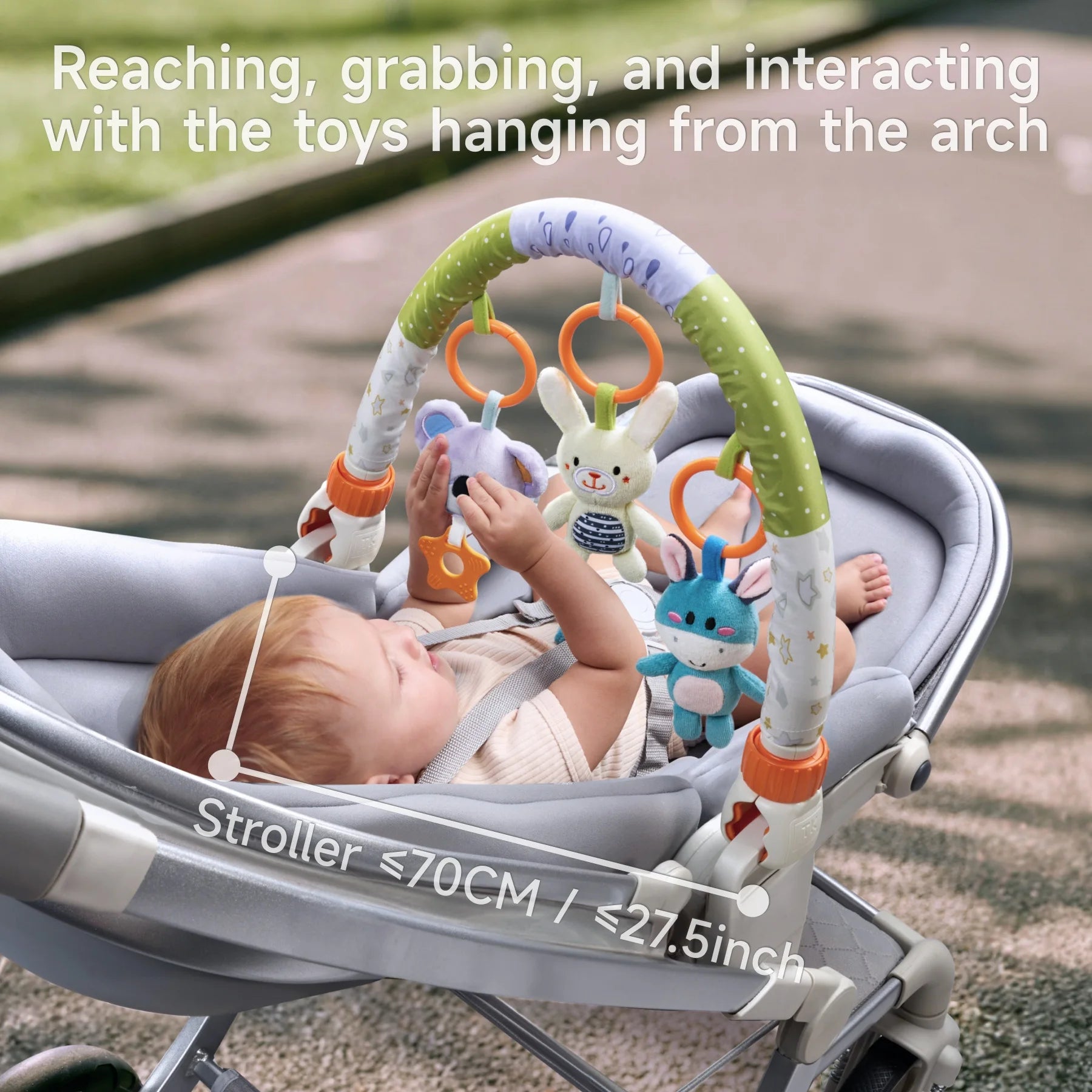 Baby lying in a stroller under a toy arch, reaching and grabbing soft animal toys hanging from the arch; caption notes stroller width ≤70cm (≤27.5 inches).