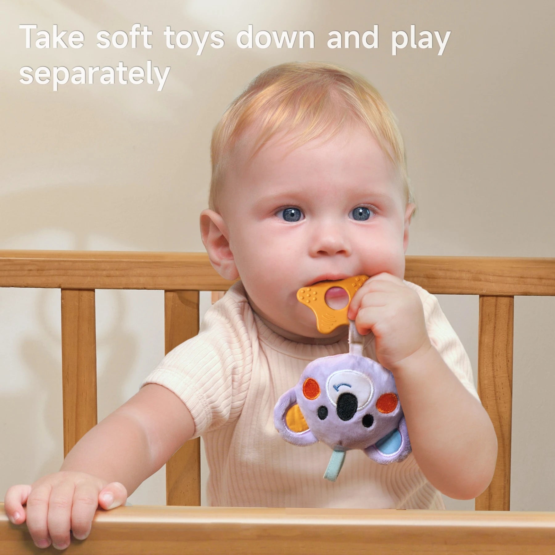 Baby standing in a wooden crib, holding and chewing on a detachable soft koala toy with a teether; caption suggests toys can be removed and played with separately.
