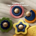 Close-up view of four colorful baby rattles designed for hanging on a play gym. The rattles are shaped like a pink circle, green flower, yellow star, and orange crescent moon, each with a smiling face in the center. The surface is textured with small embossed patterns. 
