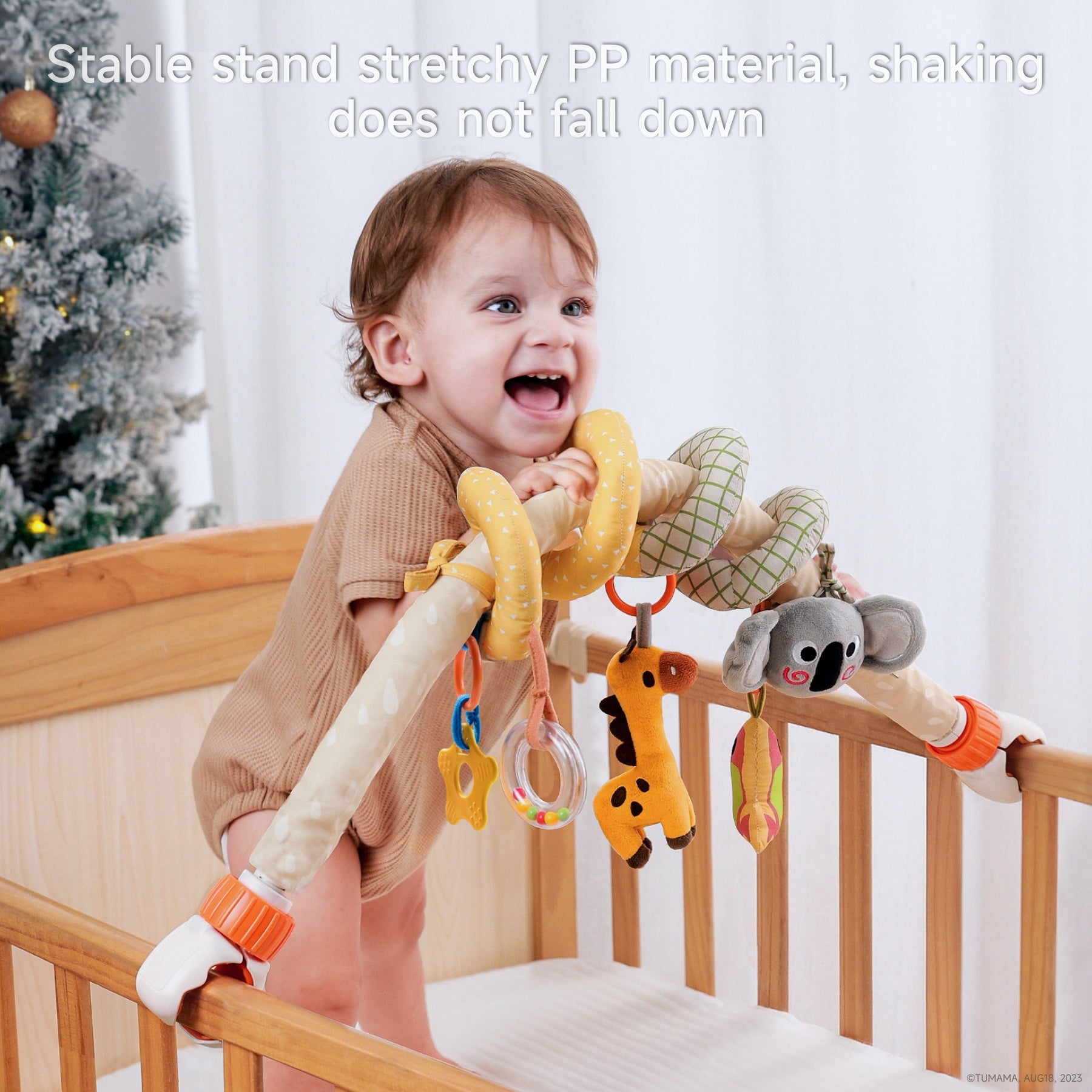Smiling toddler standing in crib holding onto a plush koala-themed spiral arch toy with hanging giraffe, parrot, teether, and rattle. Caption reads: “Stable stand stretchy PP material, shaking does not fall down.”