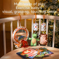 A variety of colorful baby toys including a soft mirror with plush animals, a green alligator, a pink hedgehog, and hanging cylindrical rattles attached to a crib rail. Several baby cloth books are spread out on the mattress, emphasizing play that supports visual, grasping, and touching development.