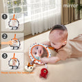 A baby lies on their tummy on a soft surface, gazing into a round plush mirror with colorful animal prints and attached hanging plush toys. The mirror reflects the baby’s face. On the left, three smaller panels show steps for using the mirror: 1) cloudy reflection with film on, 2) removing the protective film, and 3) clear reflection of the baby’s face. Text reads “Facial features cognitive mirror” and “Please remove film before use!”