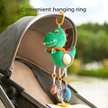 A green dinosaur plush rattle toy with a built-in mirror, teether, and sensory elements is hanging from the canopy of a stroller using an orange plastic ring. The text 