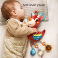 A baby peacefully sleeps on a bed while cuddling a red triceratops plush rattle toy. The soft dinosaur toy features short plush fabric and dangling sensory elements like a rattle, teether, and plush accessories. The text 