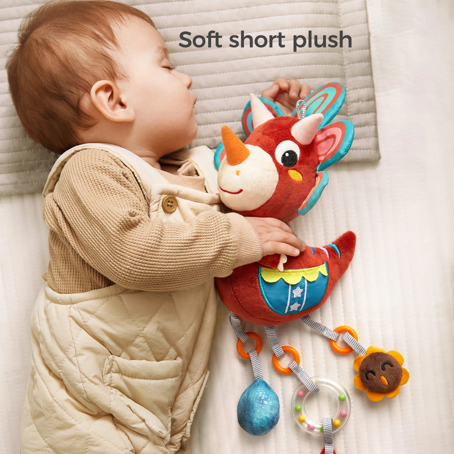 A baby peacefully sleeps on a bed while cuddling a red triceratops plush rattle toy. The soft dinosaur toy features short plush fabric and dangling sensory elements like a rattle, teether, and plush accessories. The text "Soft short plush" is displayed above, highlighting the toy’s gentle texture.