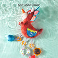 A red triceratops plush rattle toy floats on the surface of clear water, creating ripples around it. The toy features soft short plush fabric, colorful details, and attached sensory accessories including a rattle ring, teether, and plush shapes. The text 