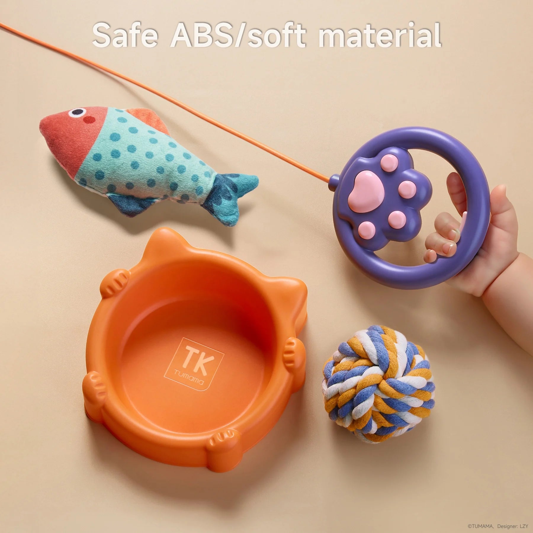 Walking cat toy close-up of toy accessories including an orange cat-shaped bowl labeled 'TK Tumama', a plush fish, a colorful rope ball, and a purple paw-shaped remote control held by a child's hand, with the caption 'Safe ABS/soft material' at the top.