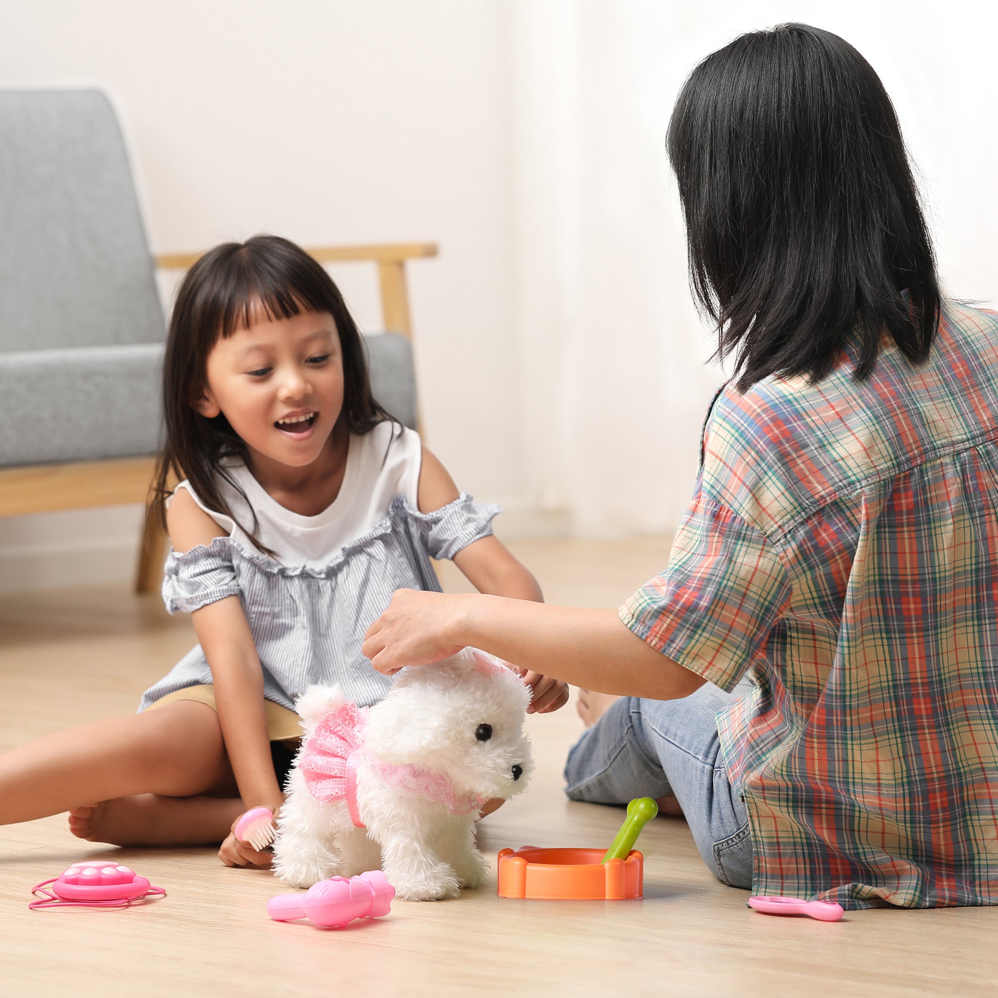 Mother and daughter sitting on the floor playing together with a white plush dog toy dressed in pink, surrounded by pretend pet grooming accessories including a bowl, bone, brush, and toy stethoscope.
