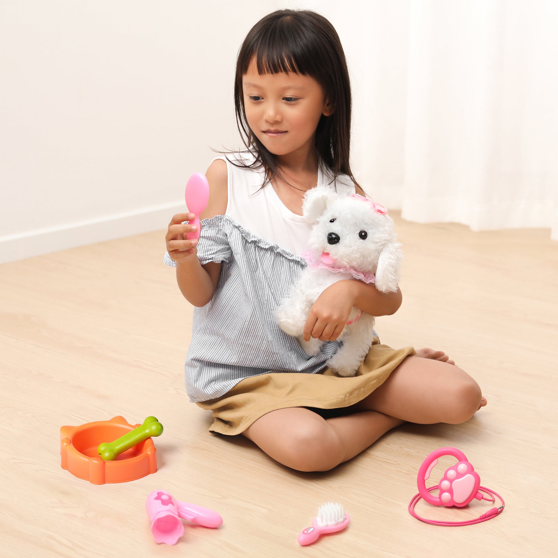 Smiling young girl sitting on the floor holding a white plush dog toy dressed in pink, while examining a toy mirror surrounded by pretend pet grooming accessories including a food bowl, bone, brush, hairdryer, and stethoscope.